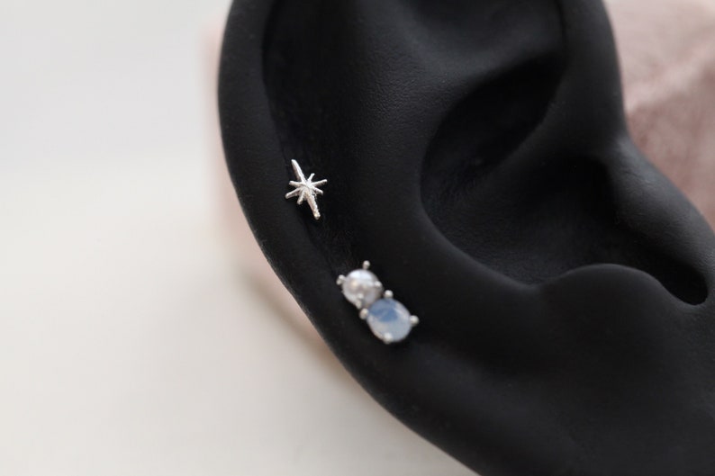 May include: A close-up of an ear with two silver earrings. The top earring is a starburst design. The lower earring has a white and blue stone. The ear is set against a black background.