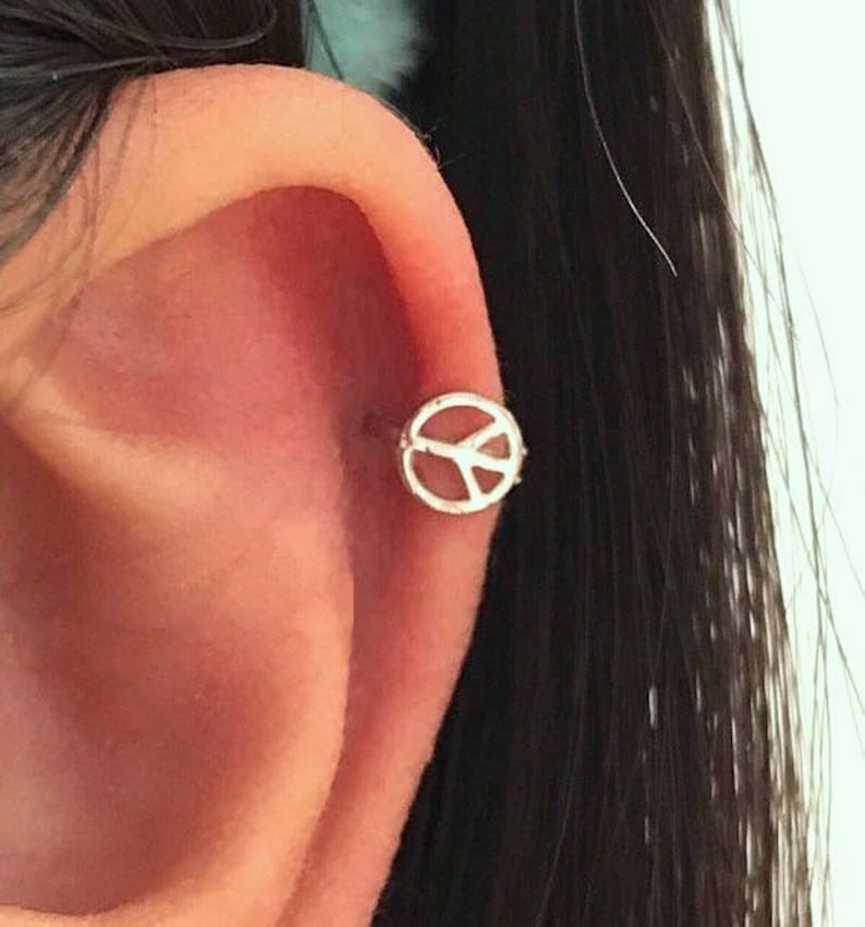 May include: A silver peace sign earring is shown on an ear. The earring is a small, circular design with the peace symbol in the center. The ear is pink and the hair is black.