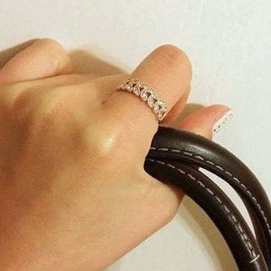 May include: A rose gold ring with a repeating leaf design is worn on a finger. The ring is set with small, clear stones. The hand is holding a black leather handbag with white stitching.