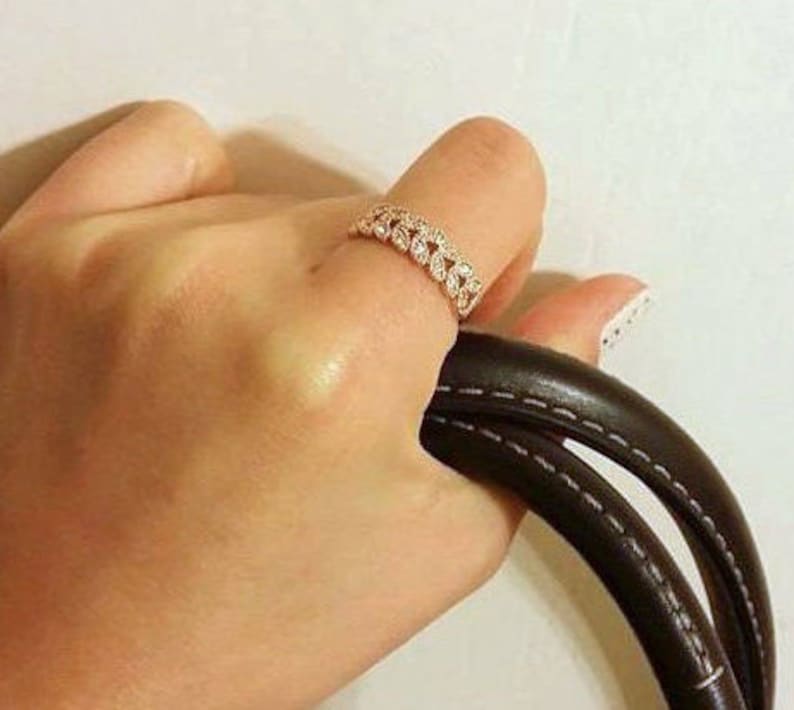May include: A rose gold ring with a repeating leaf design is worn on a finger. The ring is set with small, clear stones. The hand is holding a black leather handbag with white stitching.