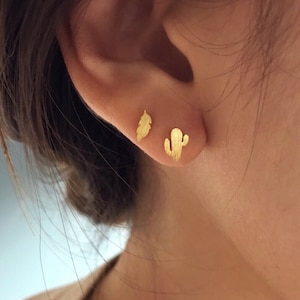 May include: A close-up of a person's ear with two gold stud earrings. One earring is a feather shape, and the other is a cactus shape. The earrings are small and delicate, with a matte finish.