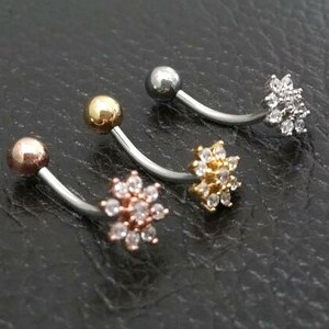 Puede incluir: Tres piercings para el ombligo curvados con diferentes acabados. Uno tiene una bola de oro rosa y una flor de cristales transparentes. Otro tiene una bola dorada y una flor de cristales transparentes. El tercero tiene una bola plateada y una flor de cristales transparentes.