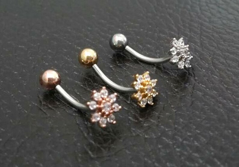 Puede incluir: Tres piercings para el ombligo curvados con diferentes acabados. Uno tiene una bola de oro rosa y una flor de cristales transparentes. Otro tiene una bola dorada y una flor de cristales transparentes. El tercero tiene una bola plateada y una flor de cristales transparentes.