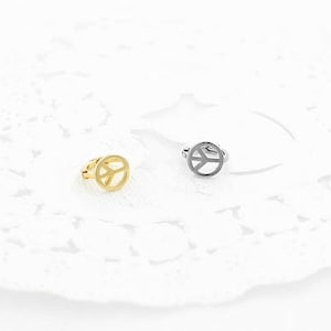 May include: Two peace sign rings. One ring is gold-colored, and the other is silver-colored. The peace sign is a circle with three lines inside, representing peace. The rings are on a white, lacy surface.