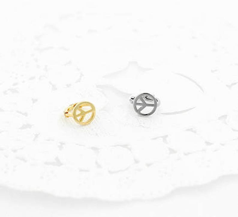 May include: Two peace sign rings. One ring is gold-colored, and the other is silver-colored. The peace sign is a circle with three lines inside, representing peace. The rings are on a white, lacy surface.