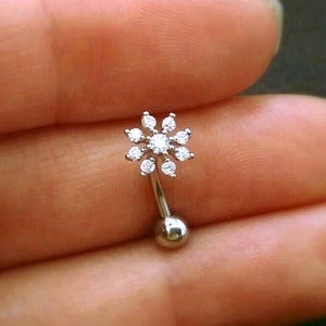Puede incluir: Un piercing de ceja curvo, de color plateado, con un dise&ntilde;o en forma de flor. La flor est&aacute; compuesta por peque&ntilde;as piedras preciosas transparentes. El piercing tiene una peque&ntilde;a bola en la parte inferior y se sostiene en una mano.