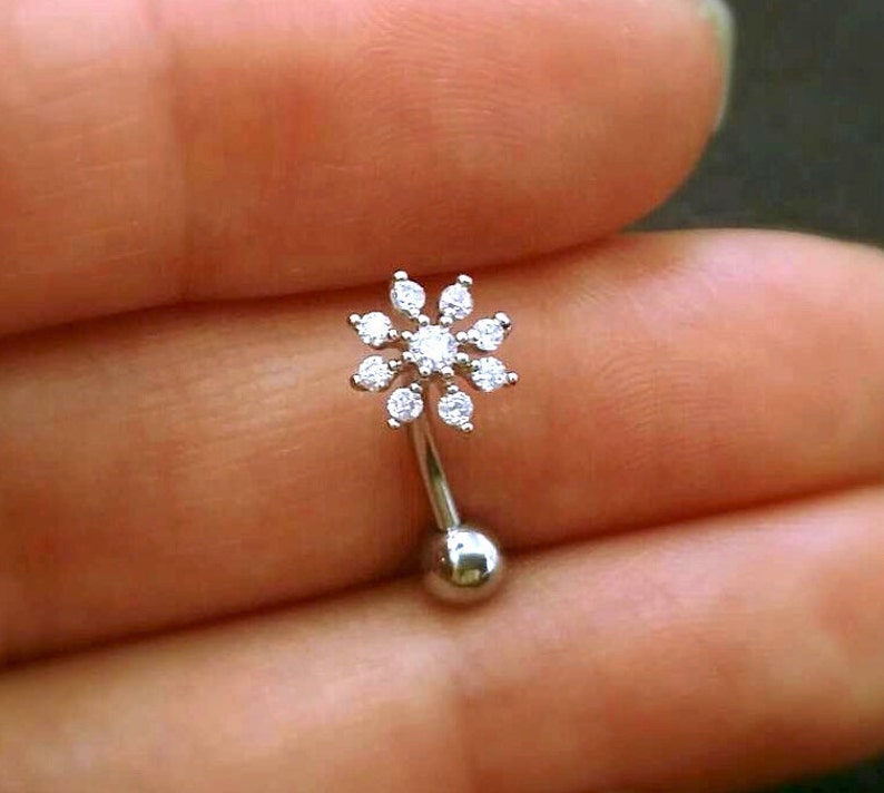 Puede incluir: Un piercing de ceja curvo, de color plateado, con un dise&ntilde;o en forma de flor. La flor est&aacute; compuesta por peque&ntilde;as piedras preciosas transparentes. El piercing tiene una peque&ntilde;a bola en la parte inferior y se sostiene en una mano.