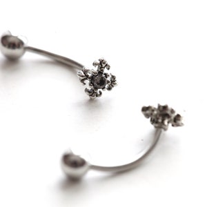 May include: Two curved, silver-toned eyebrow rings. Each ring has a heart-shaped end and a decorative, floral-like center with a dark stone. The jewelry is set against a white background.