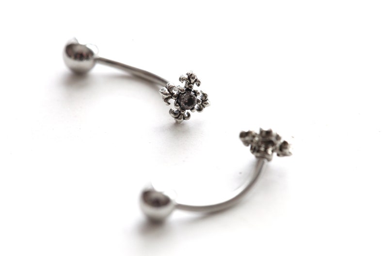 May include: Two curved, silver-toned eyebrow rings. Each ring has a heart-shaped end and a decorative, floral-like center with a dark stone. The jewelry is set against a white background.