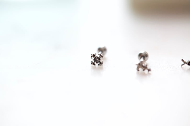 May include: Close-up of three silver-colored stud earrings. Each earring features a small, decorative, four-pointed design. The earrings are set against a bright, white background, highlighting their intricate details and metallic sheen.