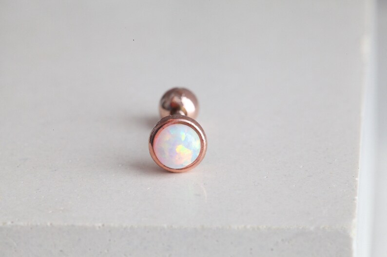 Puede incluir: Un pendiente de oro rosa con una piedra de &oacute;palo redonda e iridiscente. El pendiente tiene un poste y una peque&ntilde;a parte trasera redonda. La piedra de &oacute;palo tiene una base blanca con destellos amarillos y rosas.