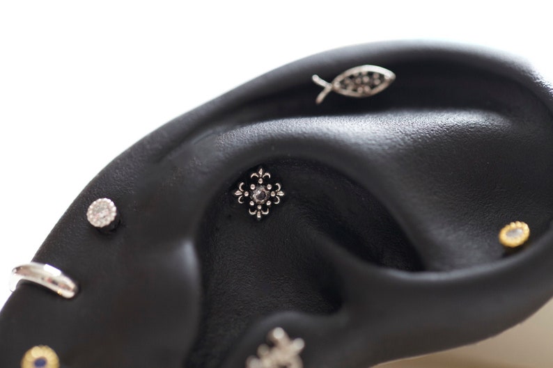 May include: Close-up of a black ear with multiple piercings. The piercings include a small silver fish, a silver stud with a clear stone, a silver stud with a flower design, and a small gold stud with a clear stone.
