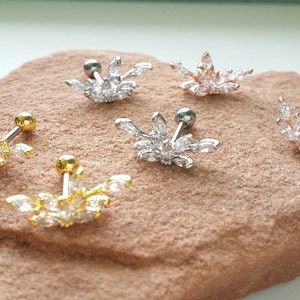 May include: An assortment of decorative ear piercings with a floral design. The piercings are in gold, silver, and rose gold tones, each adorned with clear, sparkling crystals. They are arranged on a textured, reddish-brown surface.