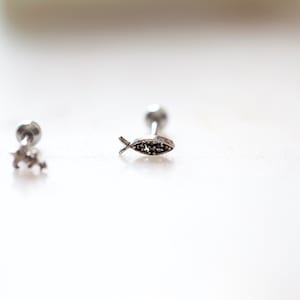May include: Close-up of three silver-colored earrings. One earring features a small fish design with dark accents. The other two earrings have a star and a round shape. The earrings are on a white surface.
