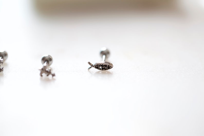 May include: Close-up of three silver-colored earrings. One earring features a small fish design with dark accents. The other two earrings have a star and a round shape. The earrings are on a white surface.