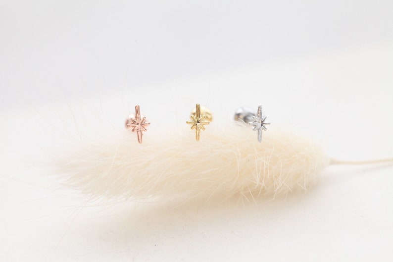 May include: Three starburst stud earrings in rose gold, gold, and silver. Each earring features a star design with multiple points. The earrings are displayed on a soft, white, feathery background.