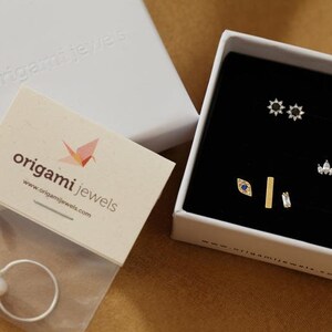May include: A white jewelry box containing several pairs of stud earrings with black and clear stones, alongside a silver ring with a white pearl in a plastic bag and a branded card with the text origami jewels and a paper crane icon.