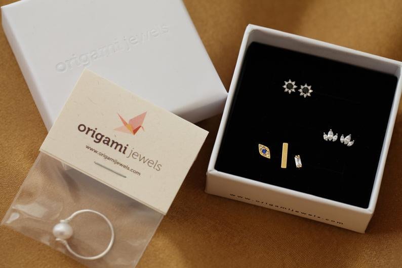 May include: A white jewelry box containing several pairs of stud earrings with black and clear stones, alongside a silver ring with a white pearl in a plastic bag and a branded card with the text origami jewels and a paper crane icon.