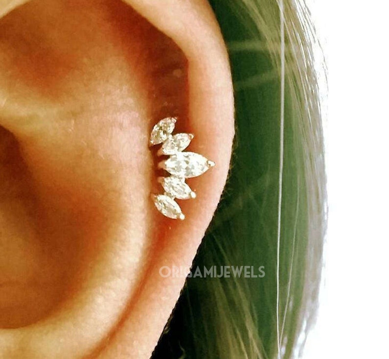 Puede incluir: Primer plano de un piercing dorado con un grupo de gemas claras con corte marquesa. Las gemas est&aacute;n dispuestas en un dise&ntilde;o curvo y en cascada. El texto "ORISAMIJEWELS" es visible en la oreja.