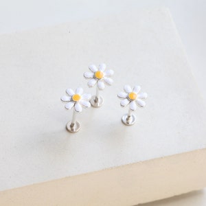 May include: Three silver-toned flat back stud earrings, each featuring a daisy flower design with white petals and a yellow center. The earrings have a straight post and a circular base, designed for body piercings with a secure flat back closure.