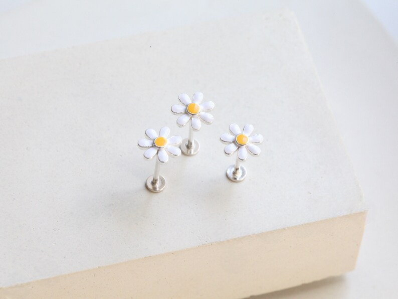 May include: Three silver-toned flat back stud earrings, each featuring a daisy flower design with white petals and a yellow center. The earrings have a straight post and a circular base, designed for body piercings with a secure flat back closure.