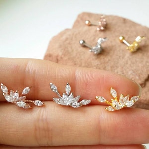 May include: Three leaf-shaped earrings with clear, marquise-cut stones are displayed. The earrings are in rose gold, silver, and gold tones. Several other stud earrings are visible in the background. The earrings are on a light background.