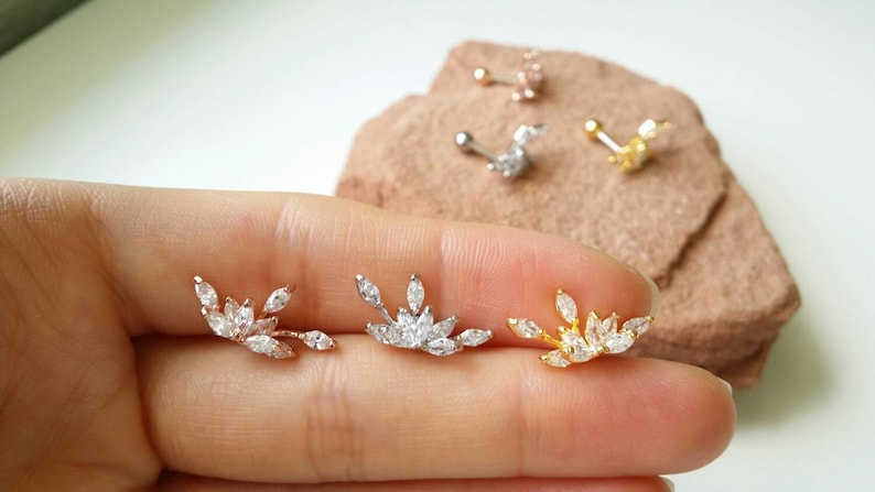 May include: Three leaf-shaped earrings with clear, marquise-cut stones are displayed. The earrings are in rose gold, silver, and gold tones. Several other stud earrings are visible in the background. The earrings are on a light background.