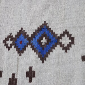 May include: A white woven rug with a geometric pattern in brown, blue, and white. The pattern features a series of connected squares with a central blue circle.