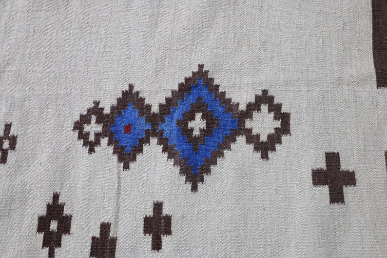 May include: A white woven rug with a geometric pattern in brown, blue, and white. The pattern features a series of connected squares with a central blue circle.