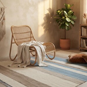 Op de afbeelding: Een lage rotan loungestoel met een gebogen rugleuning en zitting, gedrapeerd met een lichtbeige plaid. De stoel staat op een rechthoekig vloerkleed met horizontale strepen in tinten blauw, cr&egrave;me en bruin, afgewerkt met franjes aan de randen.