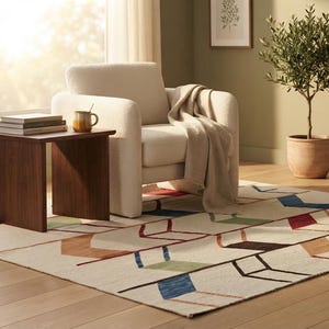 May include: A rectangular area rug featuring a cream-colored base with a geometric pattern of interconnected lines and shapes in shades of blue, green, red, and brown. The rug is positioned on a light wood floor beneath a cream armchair and table.