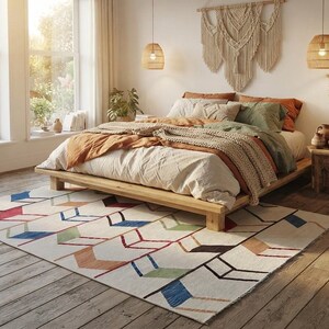 May include: A rectangular area rug featuring a repeating geometric arrow pattern in shades of blue, red, green, and brown on a cream background. The rug is positioned on a wooden floor beneath a low-profile wooden bed frame with layered bedding.