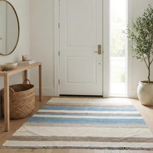 Op de afbeelding: Een rechthoekig vloerkleed met horizontale strepen in tinten lichtblauw, beige, cr&egrave;me en donkerbruin. Het kleed heeft een platgeweven textuur met korte franjes langs de randen en ligt op een lichtgekleurde houten vloer.