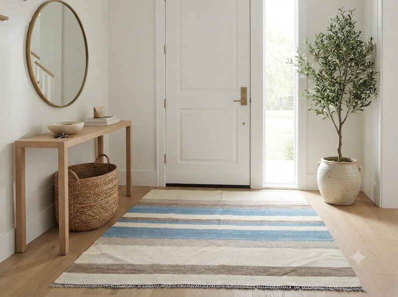 Op de afbeelding: Een rechthoekig vloerkleed met horizontale strepen in tinten lichtblauw, beige, cr&egrave;me en donkerbruin. Het kleed heeft een platgeweven textuur met korte franjes langs de randen en ligt op een lichtgekleurde houten vloer.