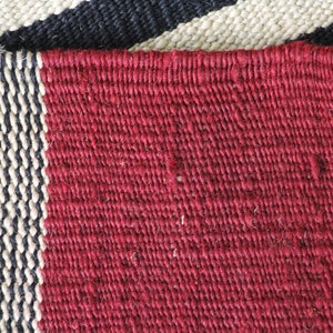 May include: Close-up of a woven rug with a red, black, and white striped pattern. The rug is made of natural fibers and has a rough texture.