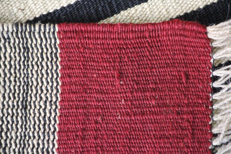 May include: Close-up of a woven rug with a red, black, and white striped pattern. The rug is made of natural fibers and has a rough texture.