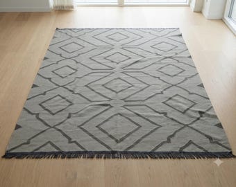 May include: A rectangular area rug featuring a light gray base with a dark gray geometric pattern consisting of interconnected diamonds and quatrefoil shapes. The rug has a short fringe along the bottom edge and a flat-woven texture throughout.