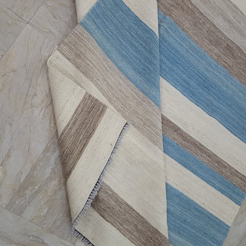 Op de afbeelding: Een geweven vloerkleed met een herhalend streeppatroon in tinten lichtblauw, beige en gebroken wit. Het kleed is omgevouwen om de onderkant te onthullen, die een vergelijkbaar streeppatroon en een korte, witte franjerand aan de hoek vertoont.
