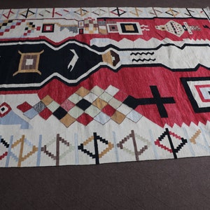 Puede incluir: Una alfombra geom&eacute;trica con patrones blancos, rojos, negros y azules con un borde de flecos. La alfombra tiene un dise&ntilde;o moderno y abstracto.