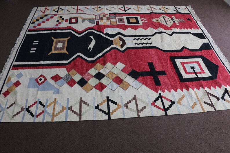 Puede incluir: Una alfombra geom&eacute;trica con patrones blancos, rojos, negros y azules con un borde de flecos. La alfombra tiene un dise&ntilde;o moderno y abstracto.