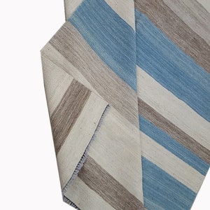 Op de afbeelding: Een geweven textiel loper met een herhalend streeppatroon in tinten lichtblauw, beige en gebroken wit. De stof vertoont een zichtbaar geweven textuur met een omgevouwen hoek die de achterkant en een afgewerkte zoomrand laat zien.