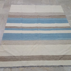 Op de afbeelding: Een rechthoekig vloerkleed met een horizontaal streeppatroon in tinten gebroken wit, lichtblauw en beige. Het geweven textiel heeft een platte textuur met zichtbare vezels en korte, gerafelde franjes langs de boven- en onderrand van het kleed.