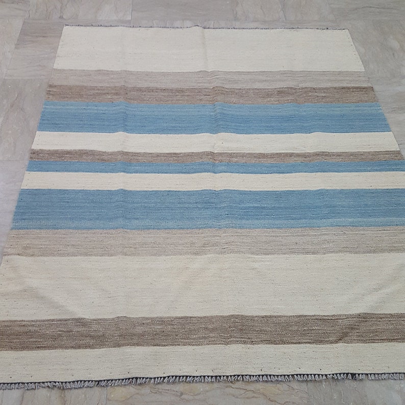 Op de afbeelding: Een rechthoekig vloerkleed met een horizontaal streeppatroon in tinten gebroken wit, lichtblauw en beige. Het geweven textiel heeft een platte textuur met zichtbare vezels en korte, gerafelde franjes langs de boven- en onderrand van het kleed.