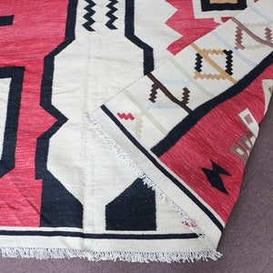 Puede incluir: Una alfombra roja, blanca y negra con estampado geom&eacute;trico y flecos. La alfombra est&aacute; doblada por la mitad y los flecos son visibles en el borde inferior.