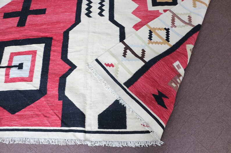 Puede incluir: Una alfombra roja, blanca y negra con estampado geom&eacute;trico y flecos. La alfombra est&aacute; doblada por la mitad y los flecos son visibles en el borde inferior.