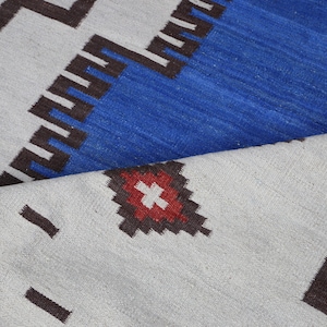 May include: A close-up of a white and blue patterned rug with a red and white diamond shape in the center. The rug has a geometric design with black lines.