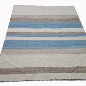 Op de afbeelding: Een rechthoekig vloerkleed met een herhalend horizontaal streeppatroon. Het ontwerp bestaat uit afwisselende banen in tinten gebroken wit, lichtblauw en gedempt bruin. Het geweven textiel heeft een plat oppervlak met een zichtbare vezelstructuur.