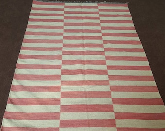 May include: A rectangular rug with a striped pattern in alternating shades of pink and off-white. The rug has a fringed edge and is laid out on a brown surface. The stripes run horizontally across the rug.