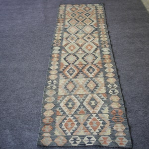 May include: A long, narrow rug with a traditional geometric pattern in shades of beige, brown, and black. The rug has a fringe on both ends.