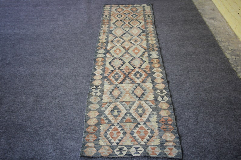 May include: A long, narrow rug with a traditional geometric pattern in shades of beige, brown, and black. The rug has a fringe on both ends.